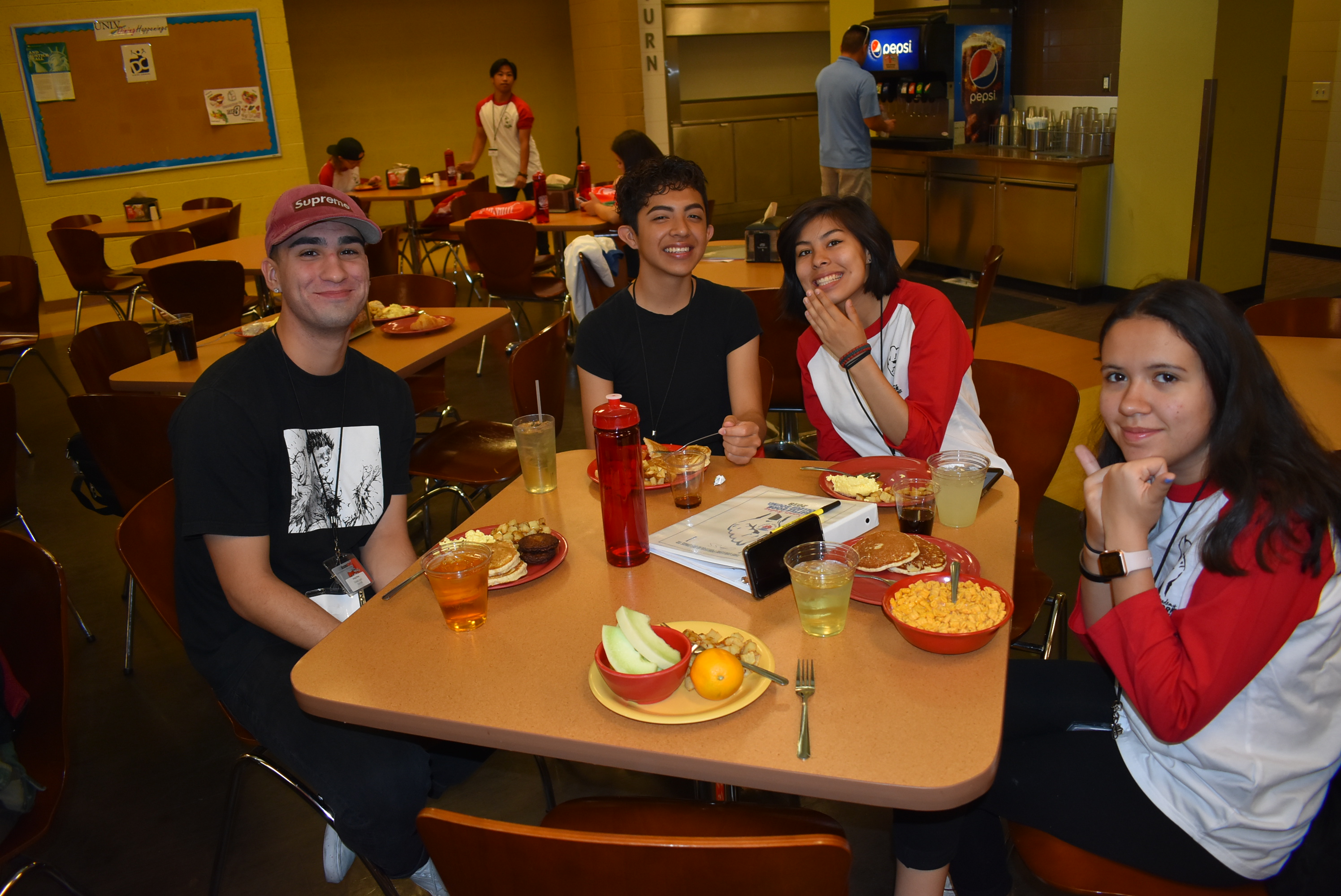 A group of young people sitting at a cafeteria table smile and pose for the camera. Two women are wearing baseball tees with red sleeves, and two men wear black shirts
