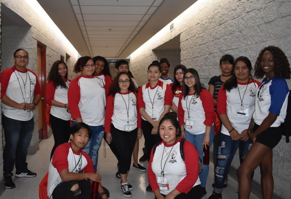 A group of students pose in for a picture inside a school hallway. Most wear baseball tees with red sleeves, while a few wear blue.