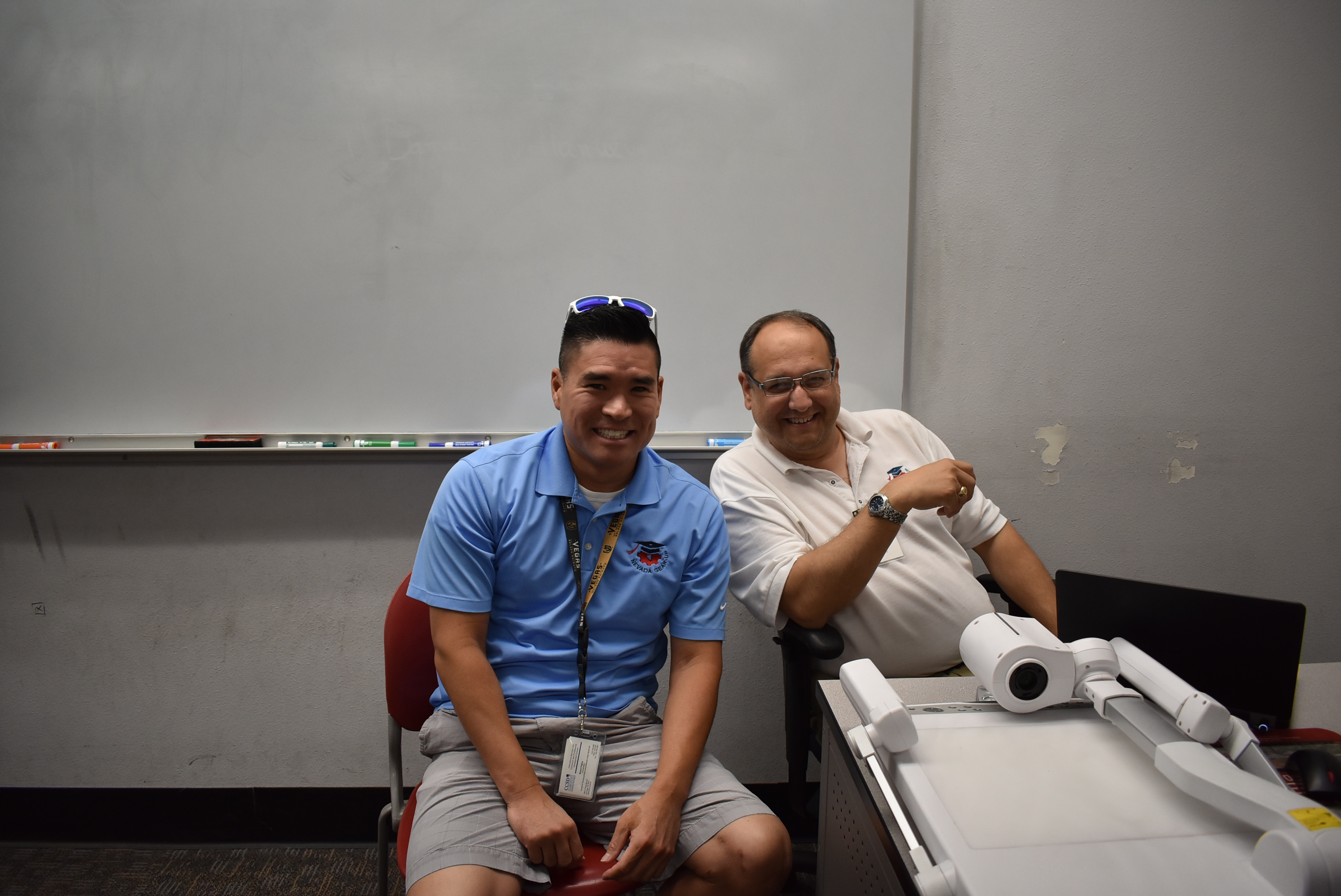 A pair of men sit near a classroom projector and smile for the camera