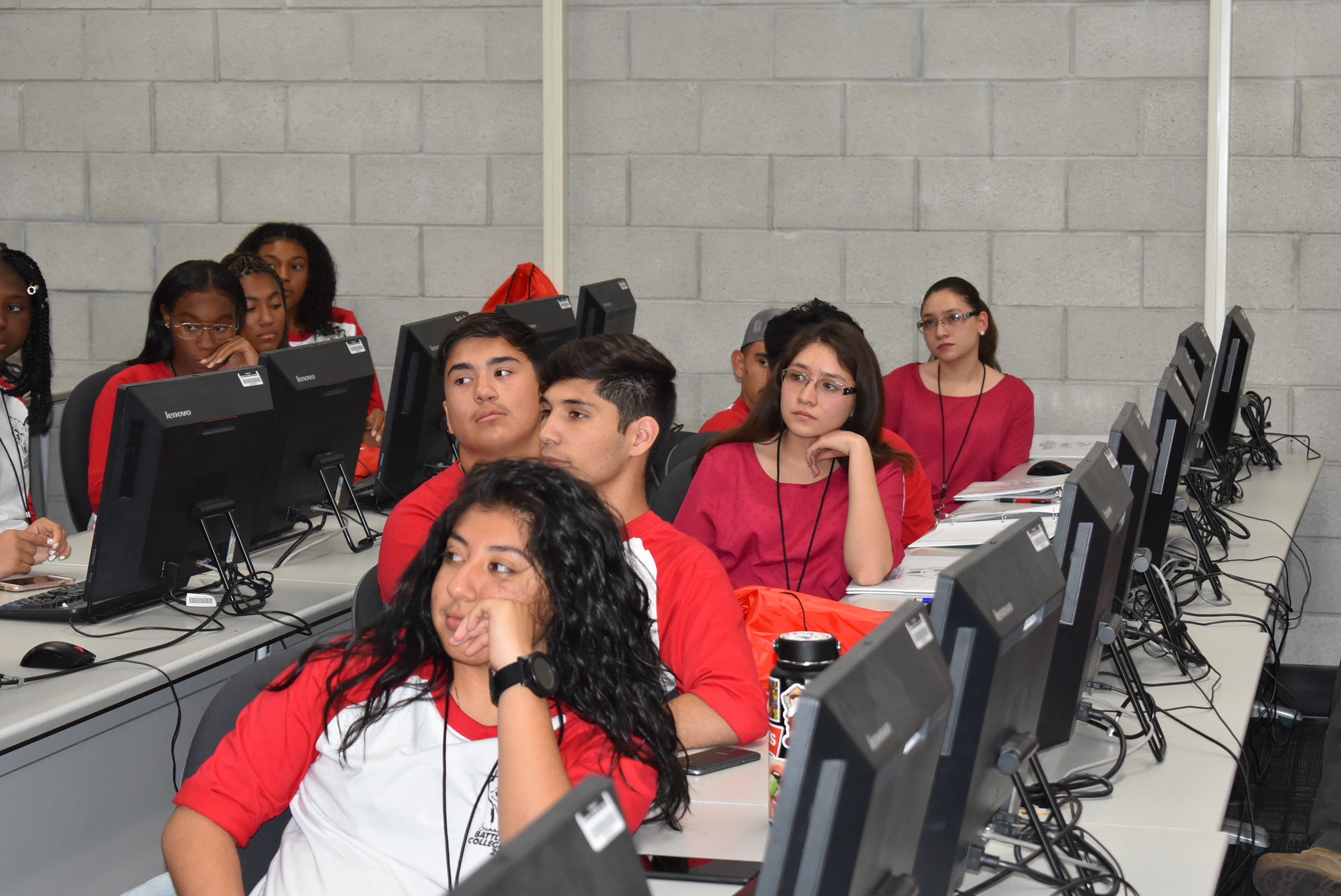 A group of students, all wearing red, sit at computer stations in a computer lab. They are all turned away from the computer monitors with their attention on a presentation.