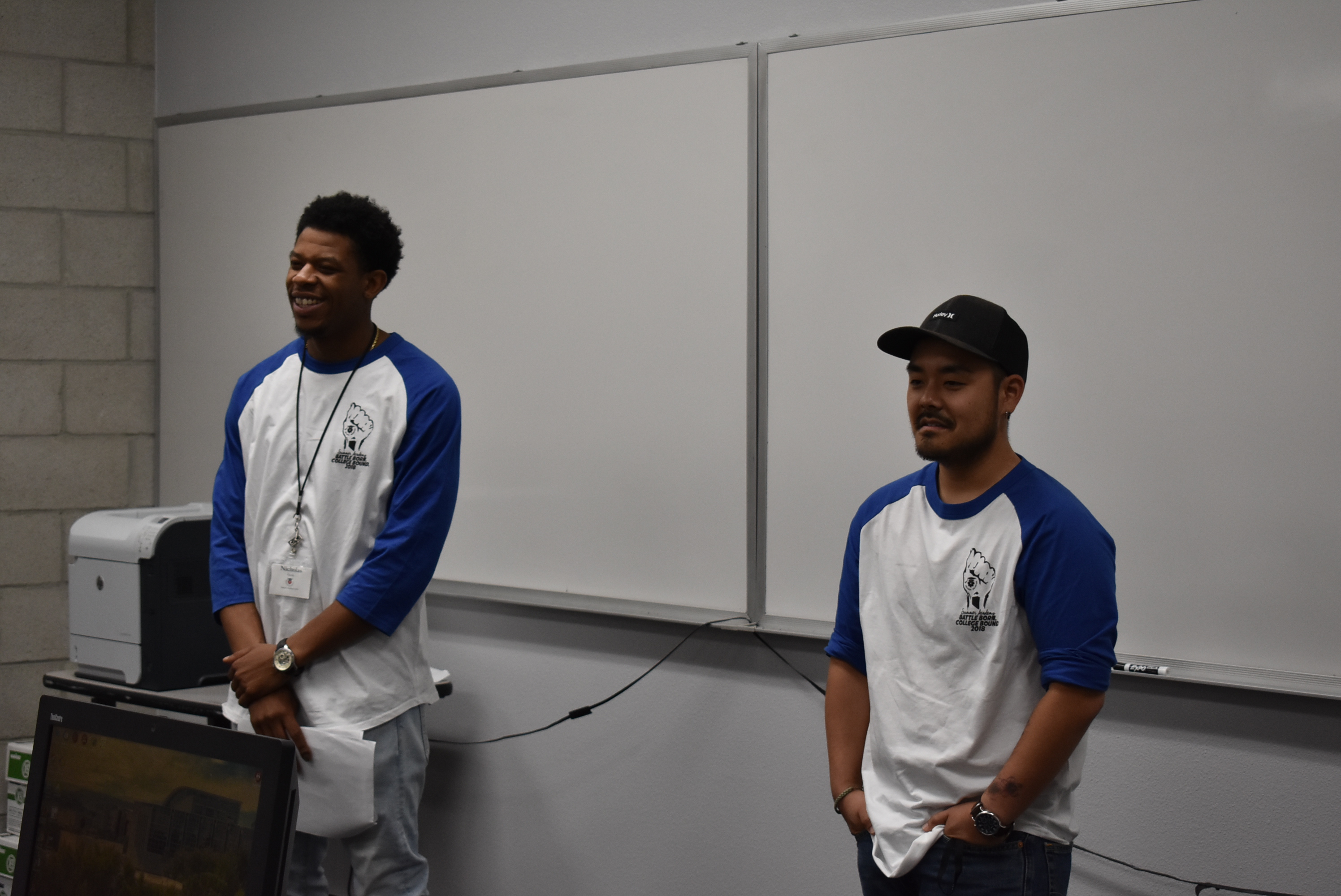 A pair of men wearing baseball tees with blue sleeves stand before a pair of blank whiteboards