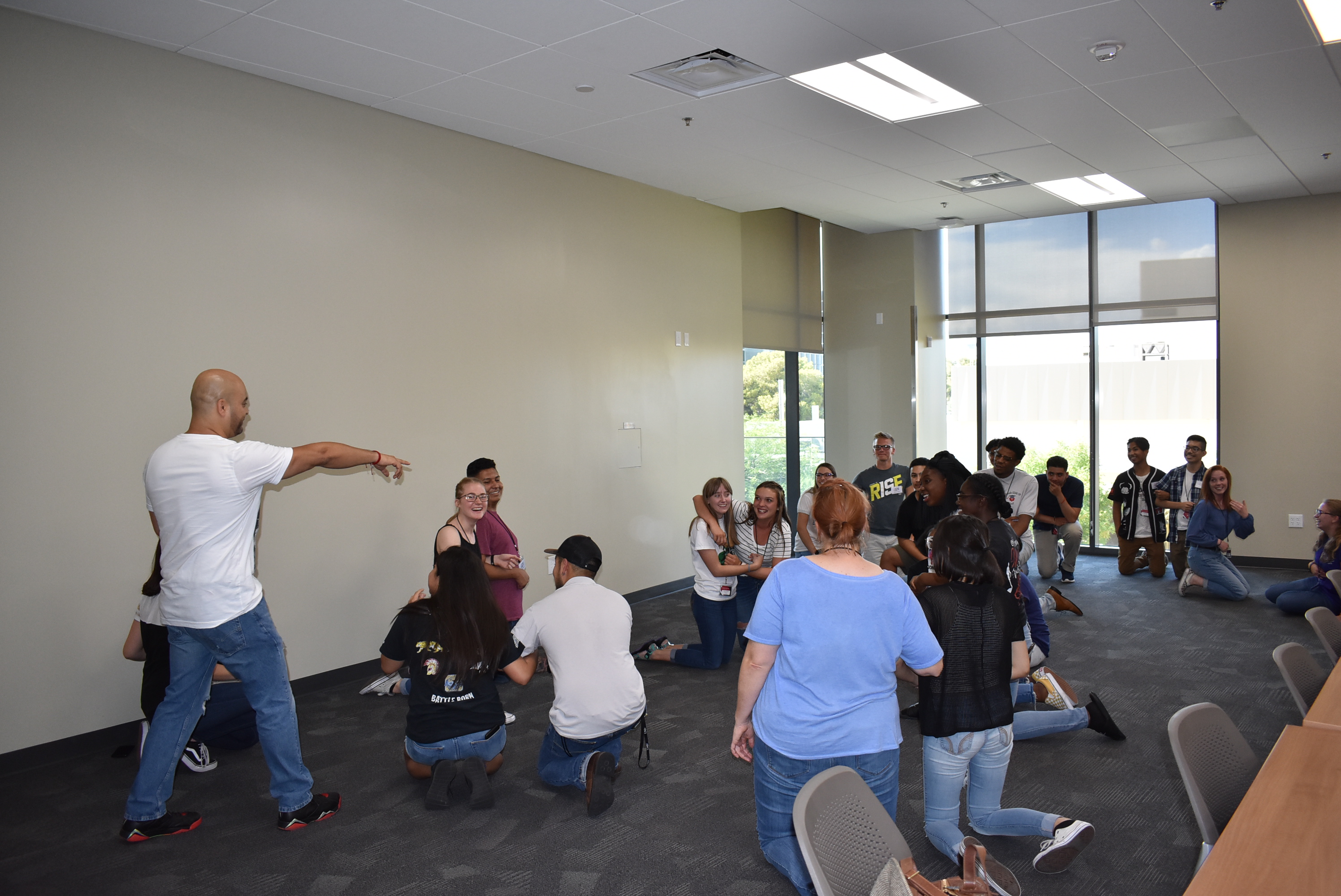 A large group of young people kneel on a classroom floor engaged in a type of teambuilding exercise. A man in a white shirt with his back to the camera appears to be leading the group through the activity.
