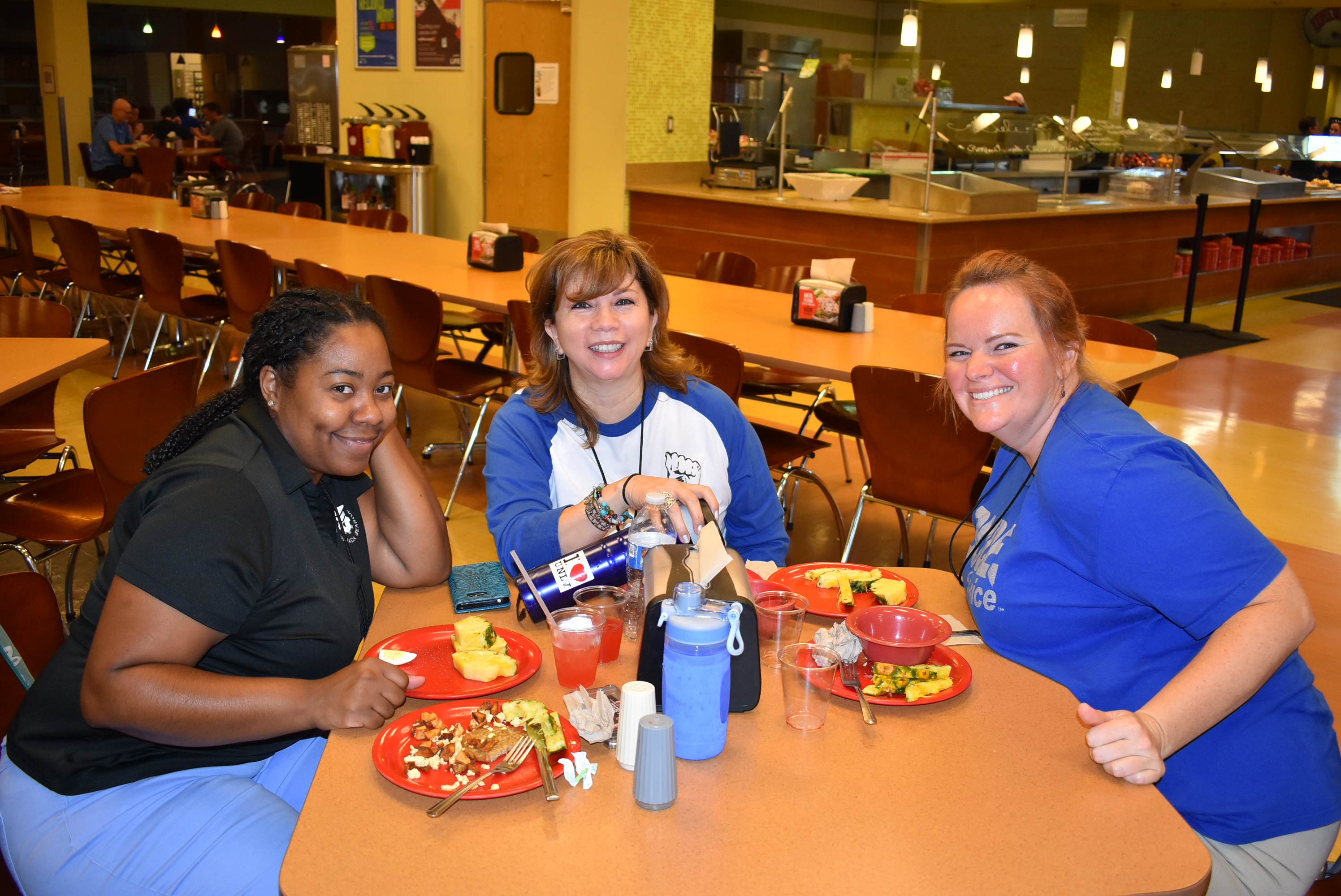 A group of three women sitting in a mostly empty cafeteria smile for the camera.