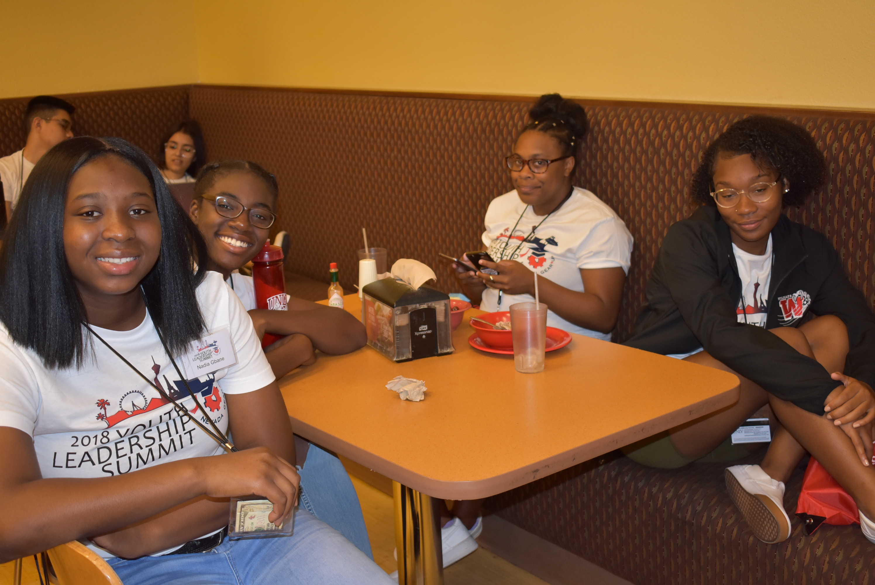 A group of four young women in matching “Youth Leadership Summit” t-shirts sit at a cafeteria table and look up to smile for the camera