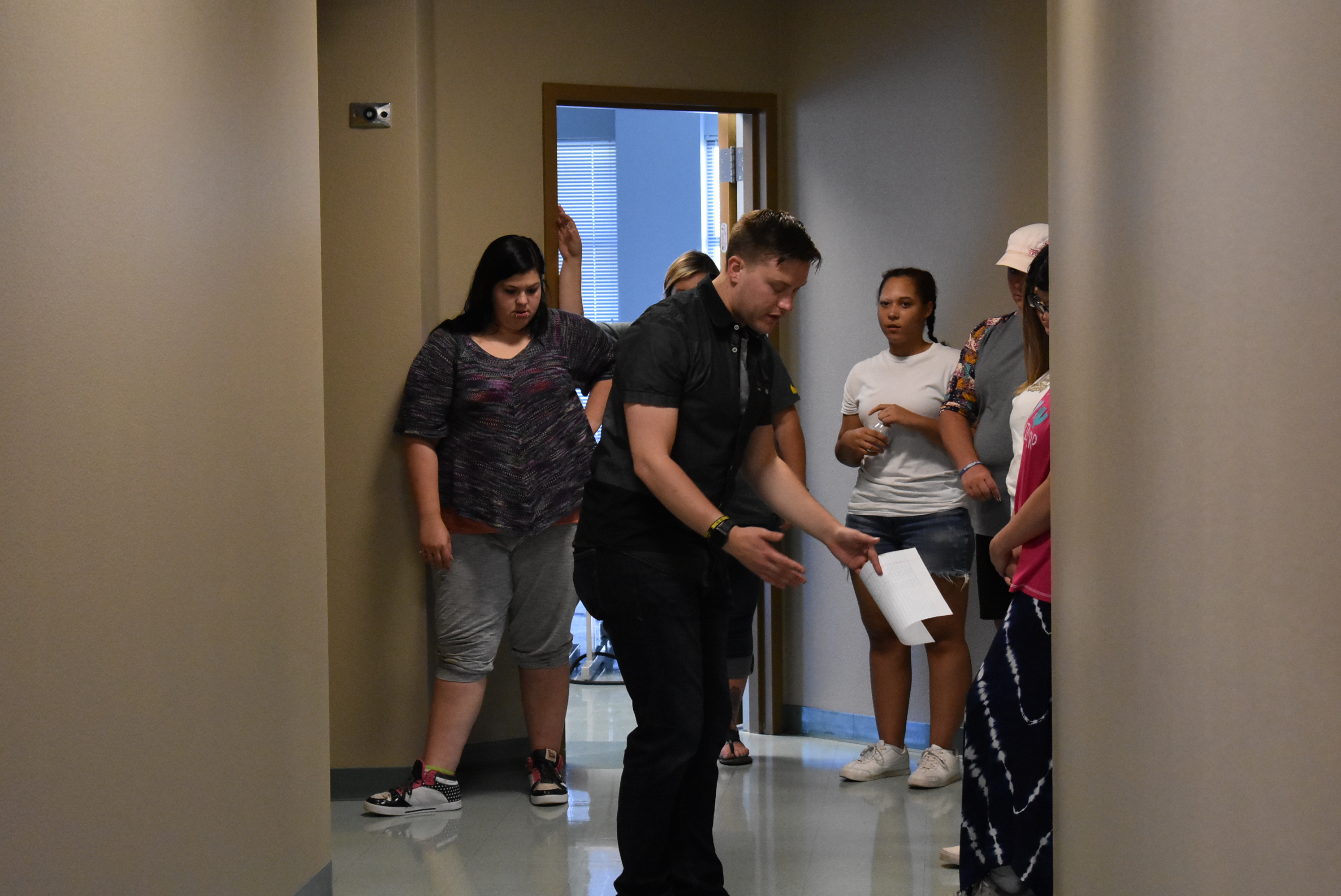 A man holding a piece of paper in one hand looks down towards a hallway floor. A group arranged in a semi-circle around the man look on.