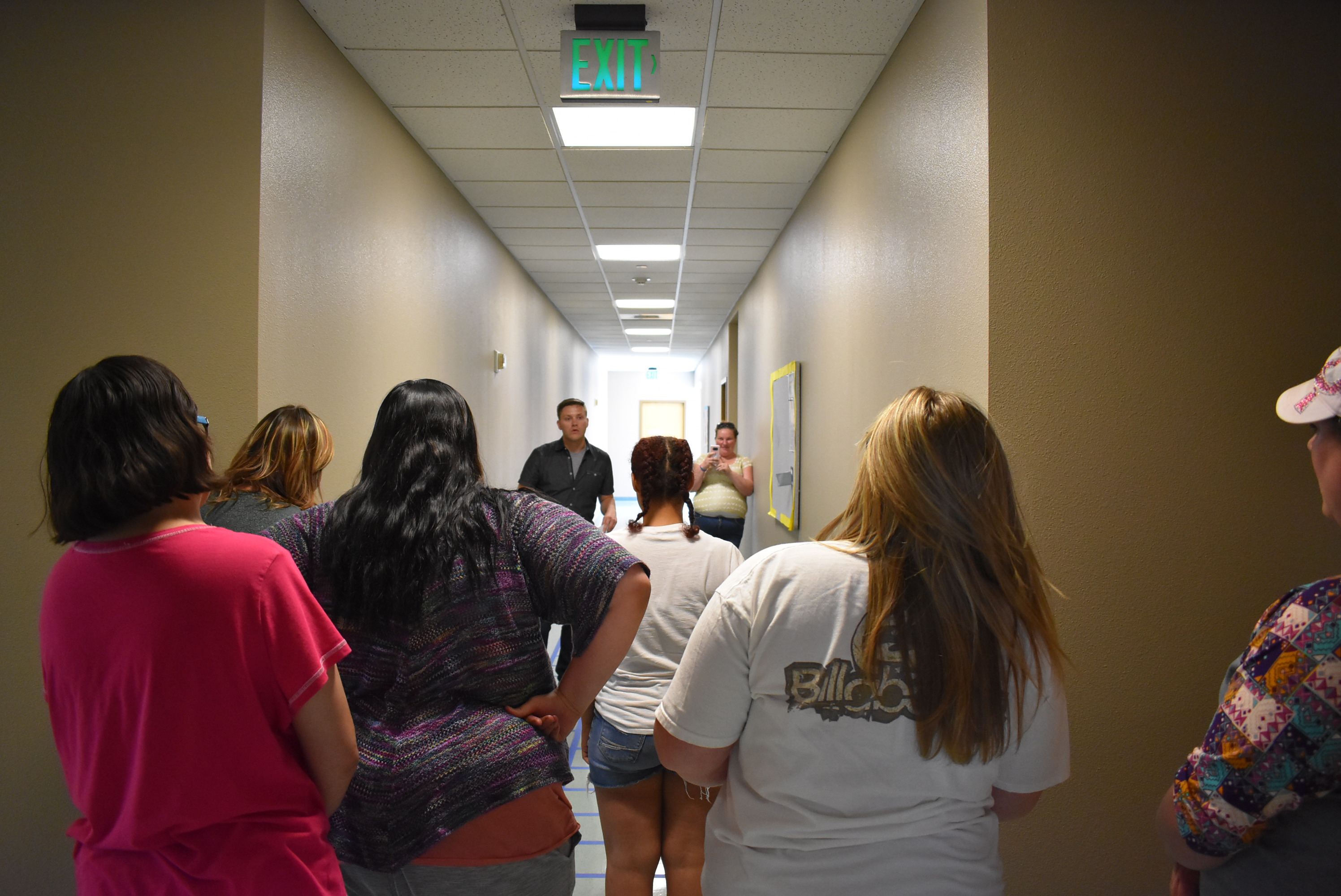 A group stands in a hallway with their backs to the camera. A man addresses this group while a woman in the background records him with her phone camera.